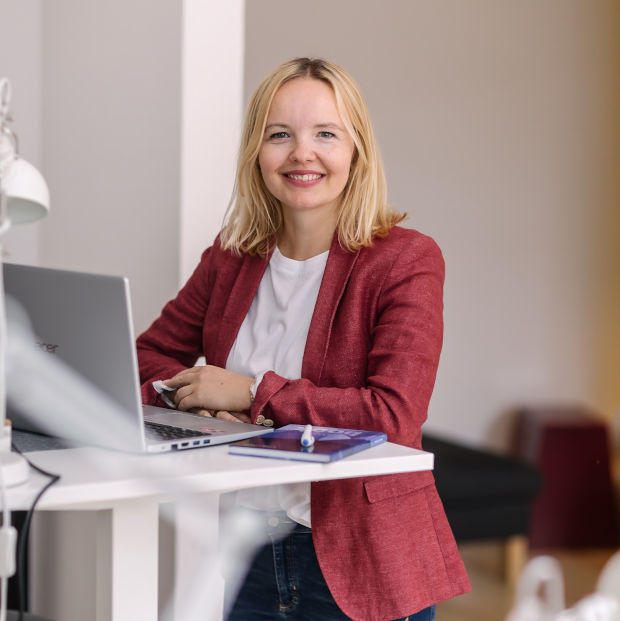 Eine lächelnde Person mit blonden Haaren sitzt an einem Tisch mit Laptop und Tablet.