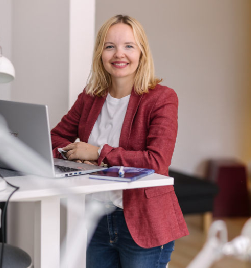 Eine lächelnde Frau in einem roten Blazer sitzt an einem Schreibtisch mit Laptop und Tablet.