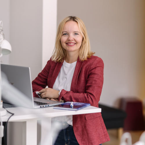 Eine lächelnde Person mit blonden Haaren sitzt an einem Tisch und arbeitet an einem Laptop.