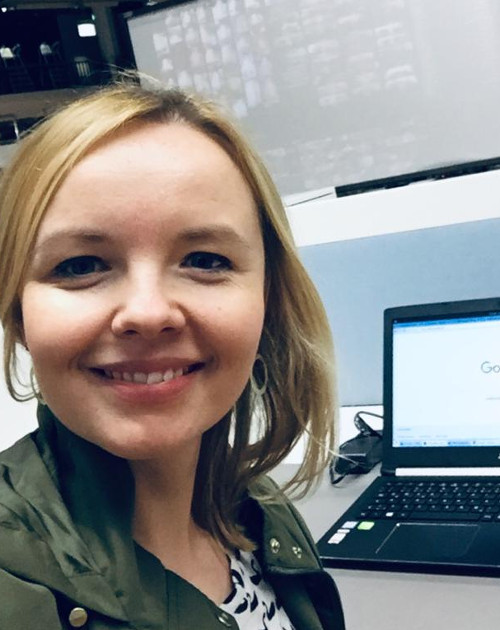 Eine lächelnde Person mit blonden Haaren steht vor einem Laptop in einem Büro.