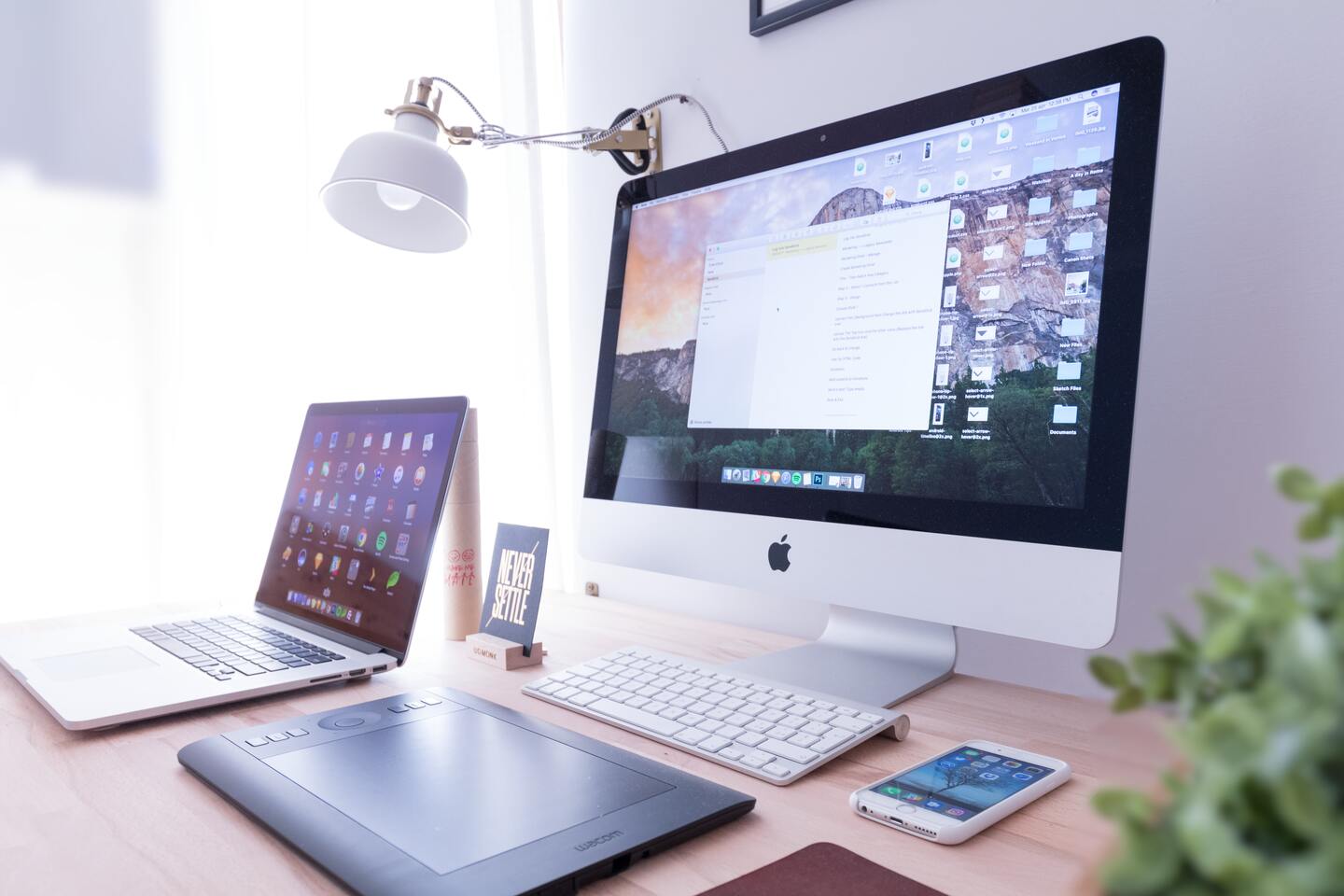 Ein gut organisiertes Arbeitsplatz-Setup mit einem iMac, einem Laptop, einer Grafiktablett, einer Tastatur, einem Smartphone und einer Pflanze.