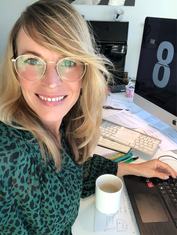 Eine Frau mit Brille sitzt lächelnd an einem Schreibtisch und tippt auf einer Tastatur, während ein Tasse Tee vor ihr steht.