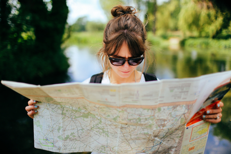 Eine Frau mit Sonnenbrille hält eine Karte und schaut darauf, im Hintergrund ist ein ruhiger See zu sehen.