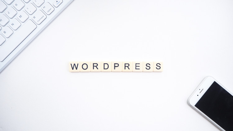 Das Bild zeigt das Wort "WORDPRESS" aus Holzbuchstaben, umgeben von einem Computer-Tastatur und einem Smartphone auf einem weißen Tisch.